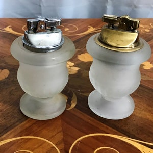 May include: Two frosted glass table lighters with silver and gold tops. The lighters are in the shape of urns with a round base.