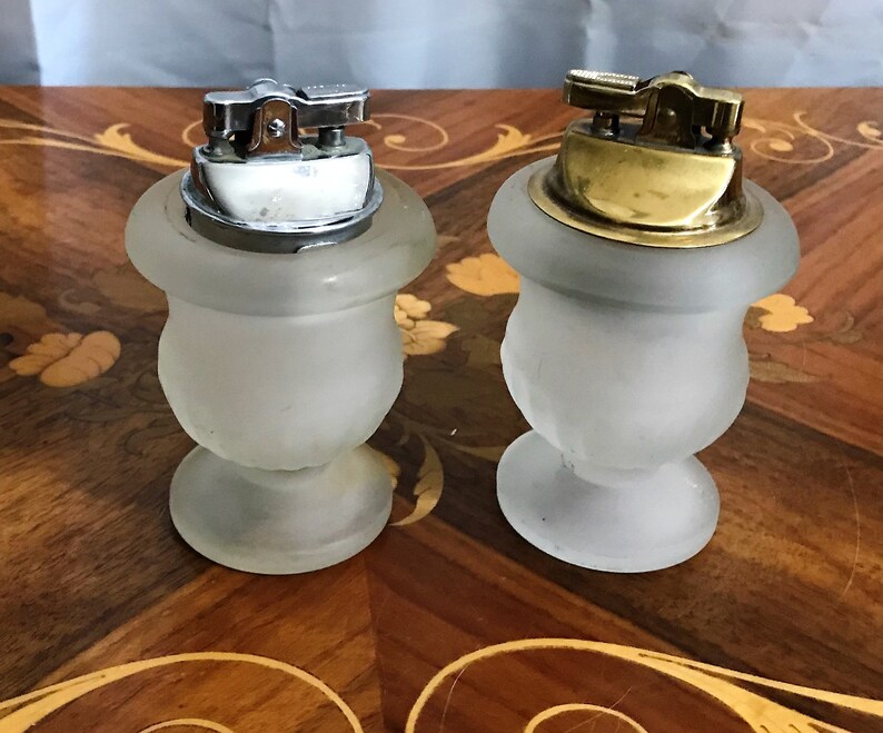 May include: Two frosted glass table lighters with silver and gold tops. The lighters are in the shape of urns with a round base.