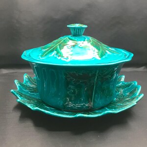 May include: A glossy turquoise ceramic tureen with a matching lid and underplate, both featuring a raised leaf pattern. The tureen has a rounded body and a fluted knob on the lid, while the underplate has a scalloped edge shaped like leaves.