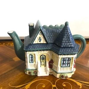May include: A ceramic teapot shaped like a house, featuring a blue roof, yellow walls, and green accents. The teapot has a chimney, a door, and windows, with red rose decorations. The spout and handle are green.