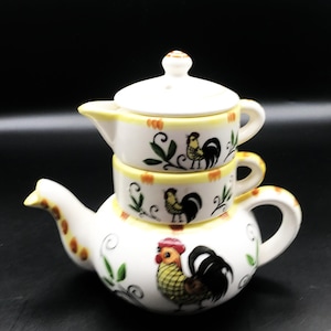 May include: A white ceramic stacking teapot set featuring hand-painted rooster motifs in yellow, red, and black. The set consists of a base teapot with a spout and handle, a middle cup, and a top lid, all decorated with green leaf accents.