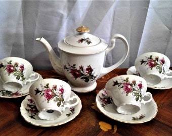 May include: A white porcelain tea set with a floral design featuring pink roses. The set includes a teapot, four cups, and four saucers.