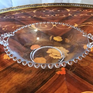 K&ouml;nnte beinhalten: Eine klare Glassch&uuml;ssel mit einem perlenbesetzten Rand und zwei Griffen. Die Sch&uuml;ssel steht auf einer Holzoberfl&auml;che mit aufwendigen Designs.