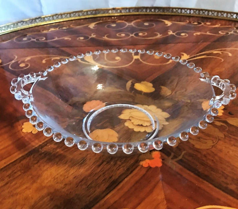 K&ouml;nnte beinhalten: Eine klare Glassch&uuml;ssel mit einem perlenbesetzten Rand und zwei Griffen. Die Sch&uuml;ssel steht auf einer Holzoberfl&auml;che mit aufwendigen Designs.