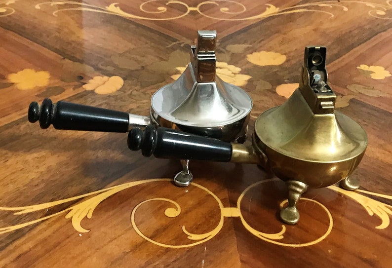 May include: Two vintage table lighters, one silver and one brass, with black handles. The lighters are on a wooden table with an intricate inlay pattern.