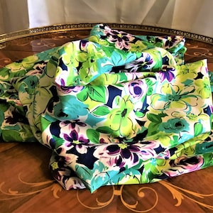 May include: A white silk scarf with a floral print in shades of green, blue, and purple. The flowers are large and bold, and the scarf is folded and draped on a wooden table.
