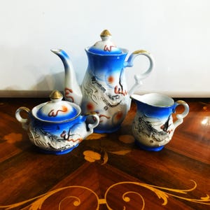 May include: A porcelain tea set featuring a teapot, sugar bowl, and creamer. The set has a blue and white gradient design with gold accents and dragon motifs. The teapot has a tall, curved spout and a lid with a gold finial.