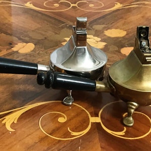 May include: Two vintage table lighters, one silver and one brass, with black handles. The lighters are on a wooden table with an intricate inlay pattern.