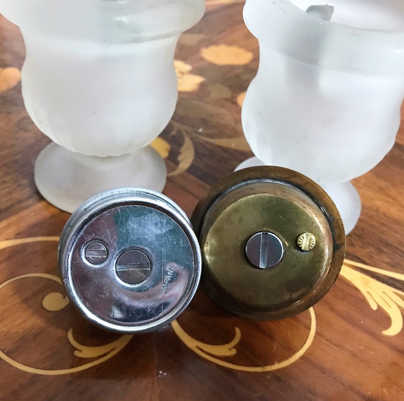 May include: Two vintage silver and brass lighters with a frosted glass base. The lighters have a round shape and a silver top with the word "Japan" printed on it. The lighters are on a wooden surface with a decorative inlay.