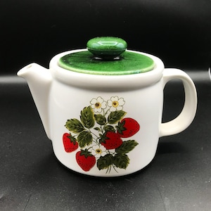May include: A white ceramic teapot featuring a vibrant botanical illustration of red strawberries, green leaves, and white flowers on the front. The teapot includes a matching removable lid with a speckled dark green glaze and a rounded handle.