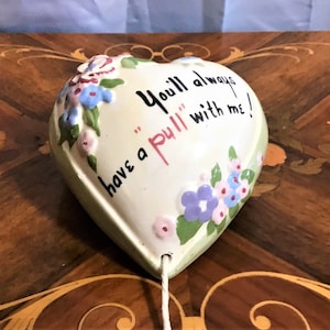 May include: A white ceramic heart-shaped ornament with green trim and painted flowers. The ornament has a string attached to the bottom and reads "You'll always have a "pull" with me!"