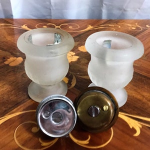 May include: Two frosted glass urns with metal lids. The urns are on a wooden surface with a decorative inlay. The lids are off and one is lying on the surface.
