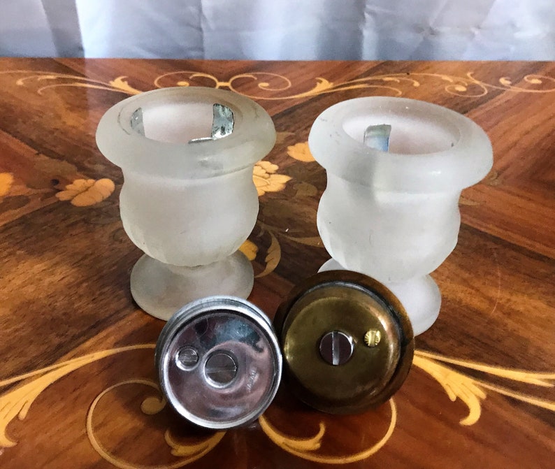 May include: Two frosted glass urns with metal lids. The urns are on a wooden surface with a decorative inlay. The lids are off and one is lying on the surface.