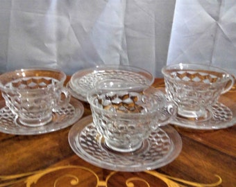 May include: Set of three clear glass teacups and saucers with a diamond pattern. The cups and saucers are arranged on a wooden table.