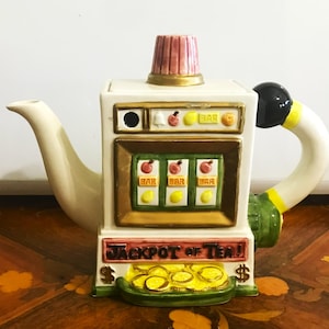 May include: A novelty teapot shaped like a slot machine, featuring a white body with a red and gold top. The teapot has a curved spout and handle, with the words "JACKPOT OF TEA!" on the front. Gold coins are at the base.