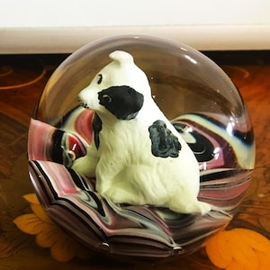May include: A clear glass paperweight featuring a white dog figurine with black spots. The dog sits atop a colorful, swirled base of pink, black, and white. The paperweight is round and has a smooth surface.