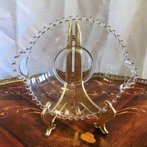 K&ouml;nnte beinhalten: Eine klare Glasservierplatte mit einem perlenbesetzten Rand und zwei Griffen. Die Platte steht auf einem goldenen Metallst&auml;nder.