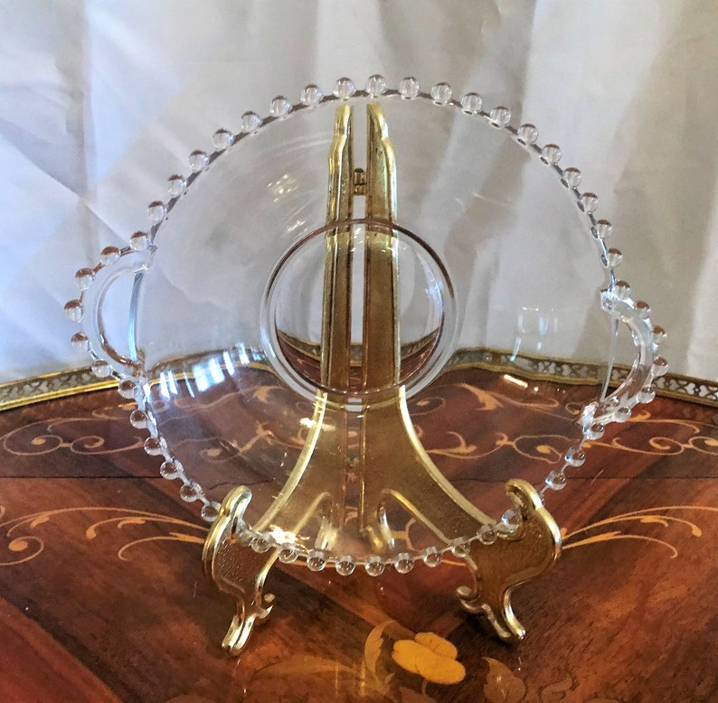 K&ouml;nnte beinhalten: Eine klare Glasservierplatte mit einem perlenbesetzten Rand und zwei Griffen. Die Platte steht auf einem goldenen Metallst&auml;nder.