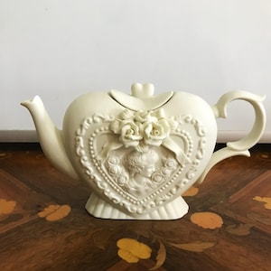 May include: Ivory-colored heart-shaped teapot with a decorative relief of a woman's profile, roses, and a ribbon. The teapot has a spout, handle, and a lid with a heart-shaped knob. The teapot is made of ceramic.