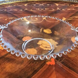 K&ouml;nnte beinhalten: Eine klare Glassch&uuml;ssel mit einem perlenbesetzten Rand und zwei Griffen. Die Sch&uuml;ssel steht auf einer Holzoberfl&auml;che mit einem Blumenmuster.