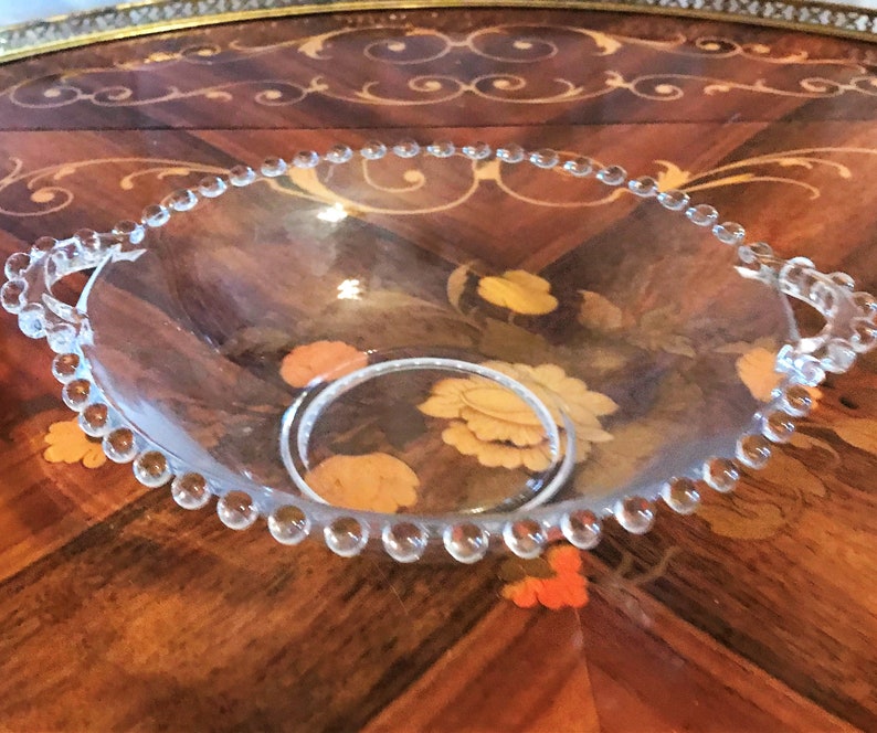K&ouml;nnte beinhalten: Eine klare Glassch&uuml;ssel mit einem perlenbesetzten Rand und zwei Griffen. Die Sch&uuml;ssel steht auf einer Holzoberfl&auml;che mit einem Blumenmuster.