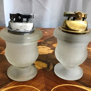 May include: Two vintage table lighters with frosted glass bases. One lighter has a silver top and the other has a gold top.