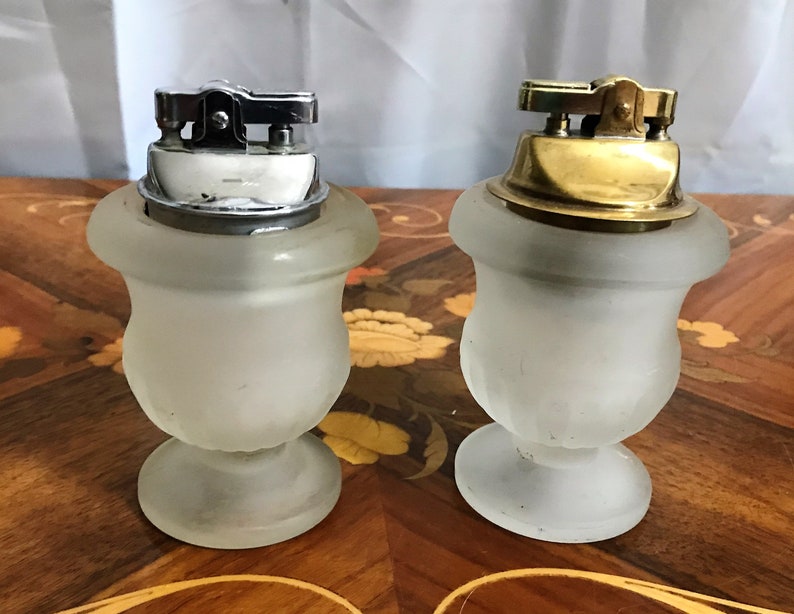 May include: Two vintage table lighters with frosted glass bases. One lighter has a silver top and the other has a gold top.