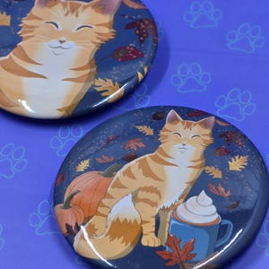 May include: Two round, blue and white enamel pins featuring a cartoon orange tabby cat with a smiling face. The first pin shows the cat sitting with a background of autumn leaves. The second pin shows the cat sitting next to a cup of coffee with whipped cream and a pumpkin.