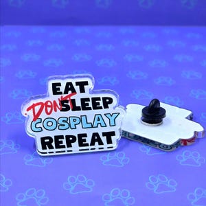 May include: A white acrylic pin with the text "Eat Don't Sleep Cosplay Repeat" in red, black, and blue lettering. The pin has a black metal backing with a safety pin.