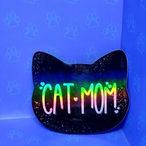 May include: A black cat head sticker with a rainbow iridescent finish. The sticker says "CAT MOM!" with hearts on either side of the text.