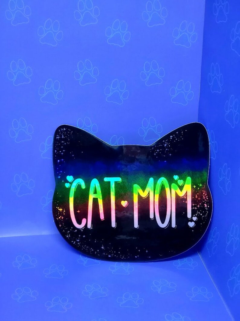 May include: A black cat head sticker with a rainbow iridescent finish. The sticker says "CAT MOM!" with hearts on either side of the text.