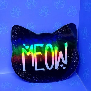 May include: A black cat-shaped sticker with a rainbow gradient and the word "MEOW!" in white text. The sticker has a glittery finish and is set against a blue background with white paw prints.