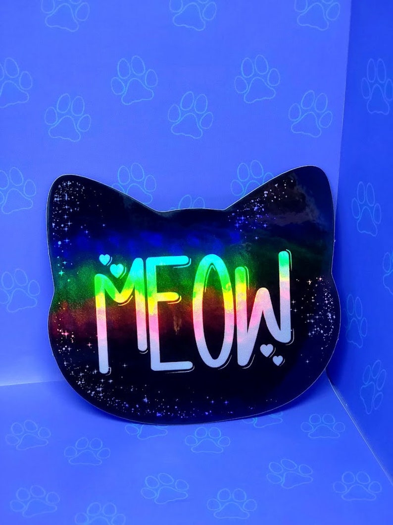 May include: A black cat-shaped sticker with a rainbow gradient and the word "MEOW!" in white text. The sticker has a glittery finish and is set against a blue background with white paw prints.