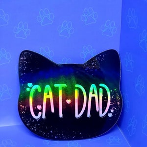 May include: A black cat head shaped sticker with a rainbow holographic finish. The sticker says "Cat Dad" with small hearts on either side of the text.