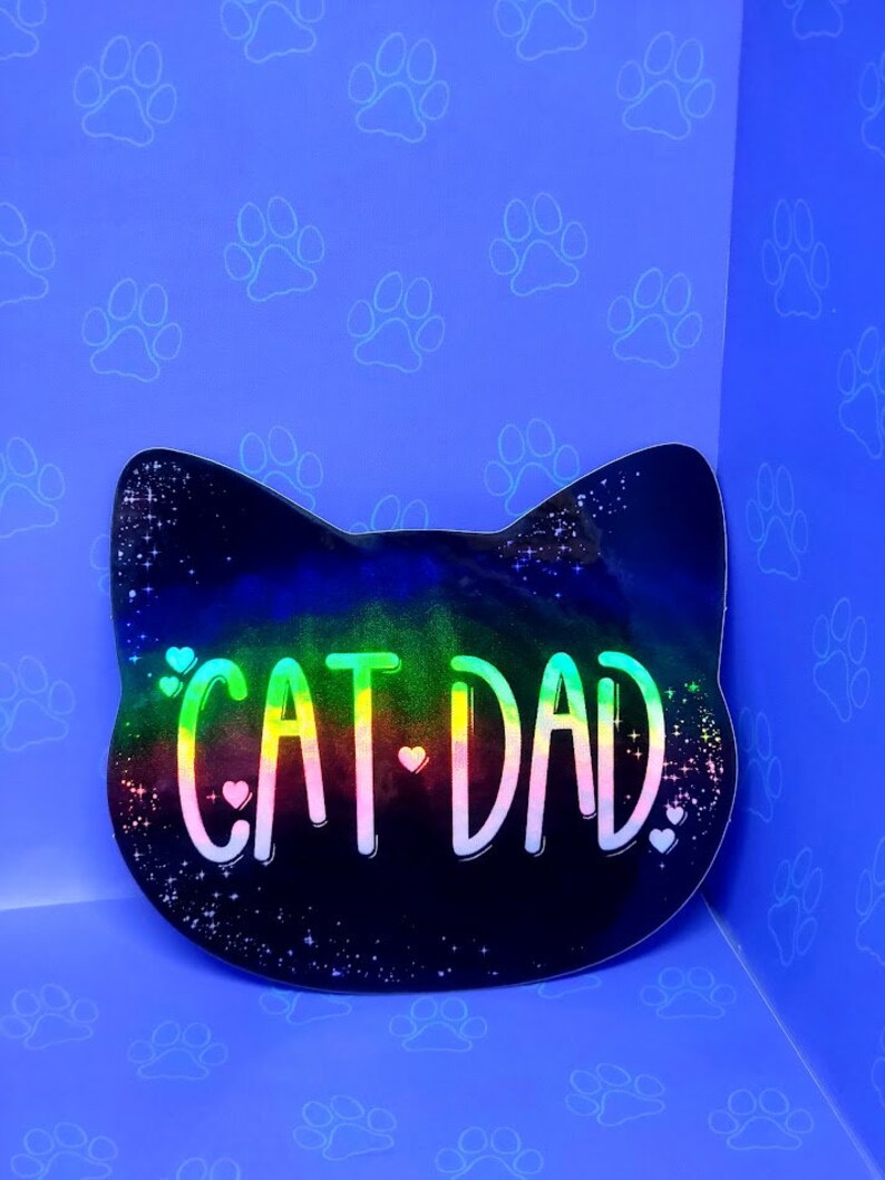May include: A black cat head shaped sticker with a rainbow holographic finish. The sticker says "Cat Dad" with small hearts on either side of the text.