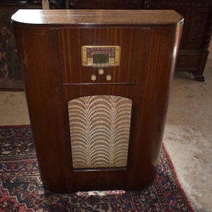 May include: A vintage wooden radio cabinet with a curved design. The cabinet is a dark brown color with a lighter brown top. It features a gold-colored control panel and a patterned speaker cover. The radio is a classic piece of furniture.