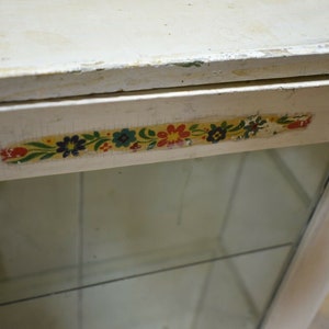 May include: White wooden cabinet with a glass door and a floral patterned trim. The cabinet has a simple design and is perfect for displaying small items.