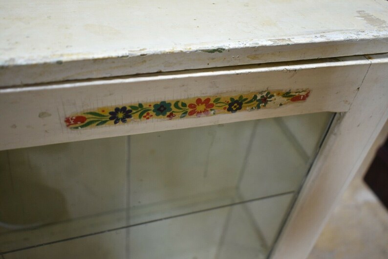 May include: White wooden cabinet with a glass door and a floral patterned trim. The cabinet has a simple design and is perfect for displaying small items.