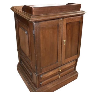 May include: A brown wooden cabinet featuring two vertical doors with a central brass keyhole, two horizontal drawers below with brass knobs, and a raised gallery edge on the top surface. The piece has a rectangular base and side panels.