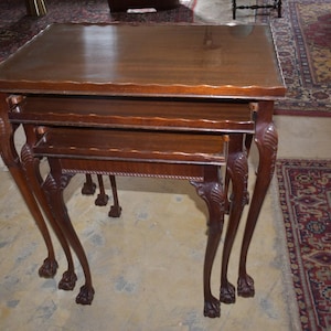 May include: A set of three nesting tables made of dark wood. The tables have a rectangular top and ornate, carved legs. The legs are curved and end in a claw-foot design. The tables are arranged in a stacked configuration.