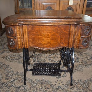 May include: A brown wooden sewing machine cabinet with a flat top surface, featuring two small drawers on each side with circular metal pulls. The cabinet is supported by a black cast iron base with a treadle pedal and a large side wheel.