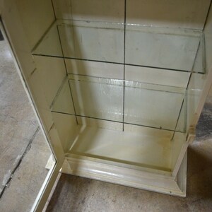 May include: A white wooden cabinet with glass shelves. The cabinet has a door that is open, revealing the empty shelves inside.