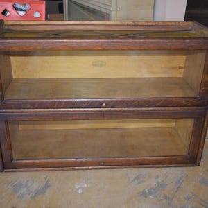 May include: A two-tiered, antique wooden bookcase with glass doors. The bookcase is made of dark-stained wood and has a rectangular shape. Each shelf has a glass door. The bookcase is designed to hold books and other items.