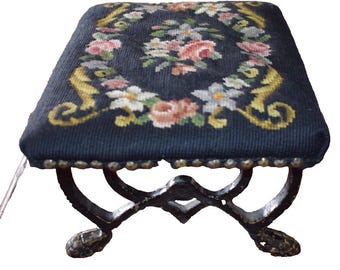 May include: An antique footstool with a dark blue needlepoint top. The top has a floral design with pink, white, and yellow accents. The legs are dark metal with a decorative, curved design. The edges of the top are lined with small, silver studs.
