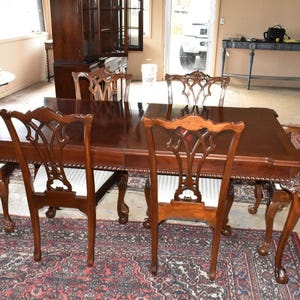 May include: A dark brown dining table with six matching chairs. The table has a polished surface and ornate legs. The chairs have white seat cushions and carved backs. The set is placed on a patterned rug, suggesting a formal dining room setting.