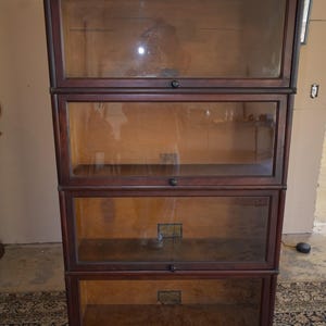 May include: A tall, dark wood barrister bookcase featuring four stacked horizontal compartments. Each section is enclosed by a clear glass door with a small, centered black knob. The interior shelves are visible through the glass, and the unit has a flat, molded top cornice and a solid base panel.