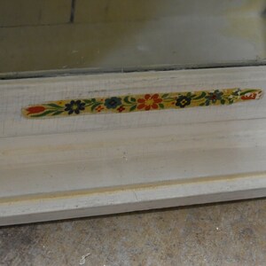 May include: A white wooden cabinet with a glass door and a floral patterned trim. The trim is yellow, green, red, and blue.