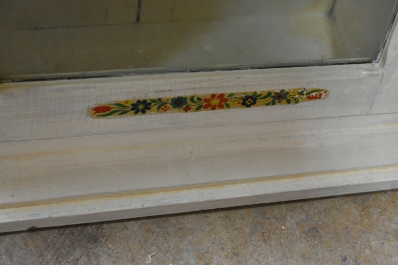 May include: A white wooden cabinet with a glass door and a floral patterned trim. The trim is yellow, green, red, and blue.