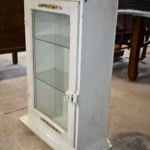 May include: White wooden cabinet with glass doors and shelves. The cabinet has a decorative trim around the edges and a small metal latch on the door.