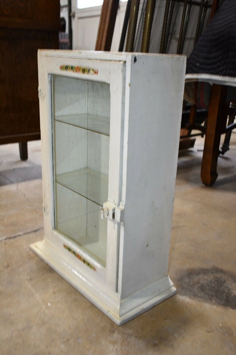 May include: White wooden cabinet with glass doors and shelves. The cabinet has a decorative trim around the edges and a small metal latch on the door.